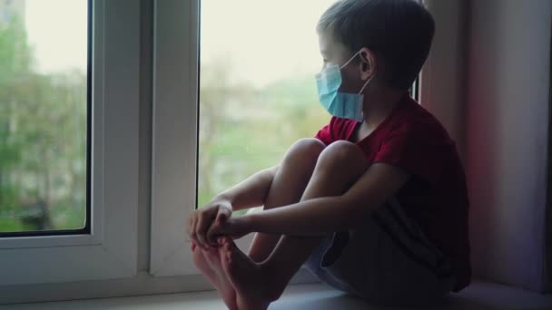 Сумний хлопчик у медичній масці сидить на підвіконні і дивиться у вікно, самокарантин. Вигляд з вулиці. Пацієнт ізольований для запобігання інфекції коронавірусу, епідемії Ковід-19, пандемії. Залишайся вдома. — стокове відео