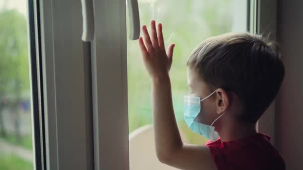 Szomorú kisfiú orvosi maszkban ül az ablak mellett és kinéz, karantén otthon, vírusvédelem — Stock videók