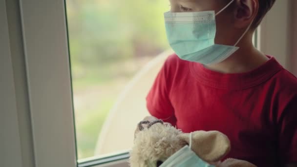 Θλιβερό παιδί στην καραντίνα. Το αγόρι και το αρκουδάκι του και οι δύο με προστατευτικές ιατρικές μάσκες κάθονται στο περβάζι του παραθύρου και κοιτάζουν έξω από το παράθυρο. Προστασία από ιούς, πανδημία του coronavirus, επιδημία πρόληψης. — Αρχείο Βίντεο