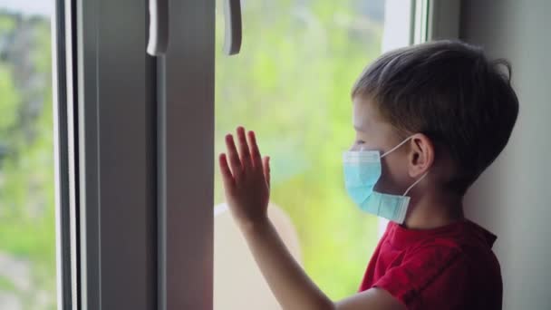 窓の近くに座って外を見る医療マスクの悲しい男の子 自宅で隔離された — ストック動画
