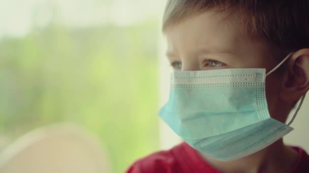 家庭内隔離の病気の子供 男の子と彼のテディベアの両方で保護医療マスクに座って窓の外を見る — ストック動画
