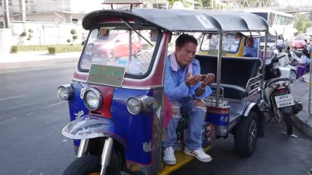 Tuk tuk driver or auto rickshaw plays on the phone while waiting for a new passenger. BANGKOK, THAILAND - DECEMBER 02, 2019 — ストック動画