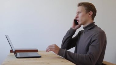 Genç adam dizüstü bilgisayar kullanıyor ve telefonda konuşuyor. Profesyonel yaratıcı milenyum, ofis stüdyosundaki masasında çalışıyor. Öğrenci çevrimiçi çalışıyor. 4k