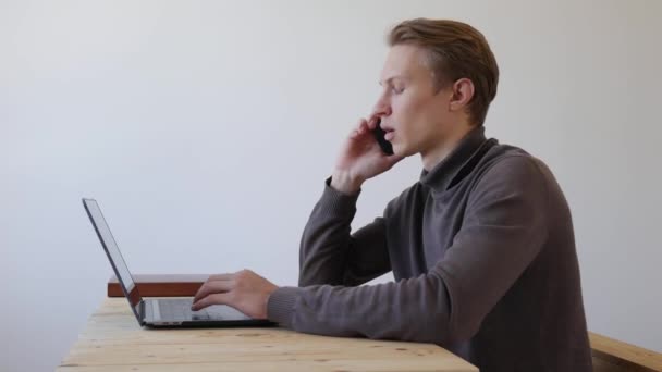Genç adam dizüstü bilgisayar kullanıyor ve telefonda konuşuyor. Profesyonel yaratıcı milenyum, ofis stüdyosundaki masasında çalışıyor. Öğrenci çevrimiçi çalışıyor. 4k — Stok video