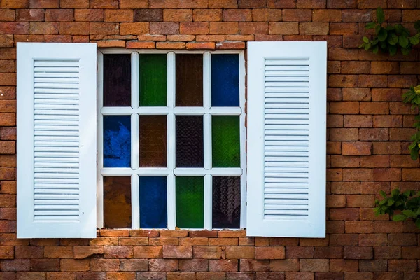 Vecchia finestra di legno bianco con vetrate su sfondo muro di mattoni marrone, muro di pietra con piccola vetrata colorata e foglia verde sul lato destro . — Foto Stock