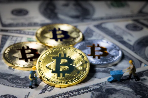 Маленький шахтёр выкапывает цифровую валюту Golden bitcoin на фоне банкнот за сто долларов. Электронные деньги, цифровые деньги, концептуальный образ добычи и торговли биткойнами . — стоковое фото