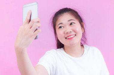 Güzel genç Asyalı kadın portresi beyaz bir tişört giy. Selfie alma ve pembe arka plan gülümseyen bir akıllı telefon kullanarak. Güzel gözlü Asyalı kadın.