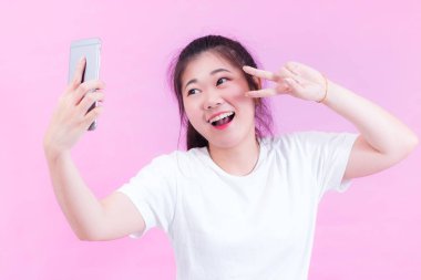 Güzel genç Asyalı kadın portresi beyaz bir tişört giy. Selfie alma ve gülümseyen ve v-sign pembe arka plan üzerinde gösterilen bir akıllı telefon kullanarak. Güzel gözlü Asyalı kadın.