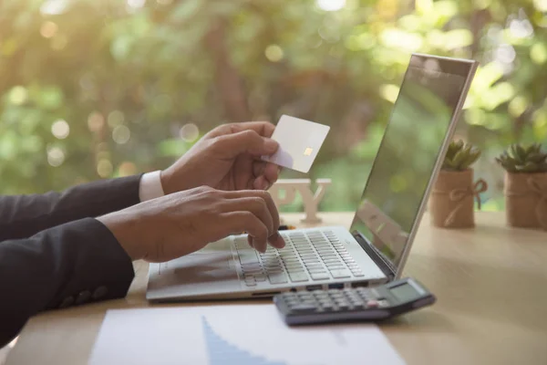 Vista posteriore di moderni uomini d'affari in possesso di carte di credito digitando numeri sulla tastiera del computer mentre seduto con la penna, calcolatrice su tavolo in legno, messa a fuoco morbida, luce del sole bagliore. Concetto di shopping online . — Foto Stock