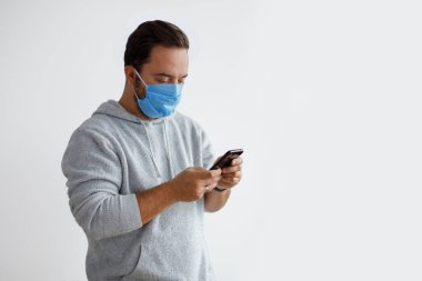 Beyaz sakallı yetişkin bir adam beyaz bir arka planda izole edilmiş cerrahi maske takarken akıllı telefon kullanıyor. Sağlık, salgın hastalıklar, sosyal medya, iletişim ve fotokopi alanı ile yaşam tarzları
