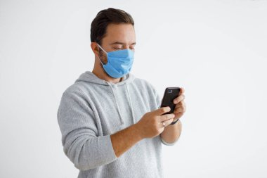 Beyaz sakallı yetişkin bir adam beyaz bir arka planda izole edilmiş cerrahi maske takarken akıllı telefon kullanıyor. Sağlık, salgın hastalıklar, sosyal medya, iletişim ve fotokopi alanı ile yaşam tarzları