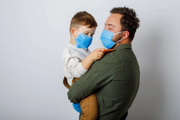 Genç babası ve küçük oğlu beyaz bir arka planda cerrahi maske takıyor.
