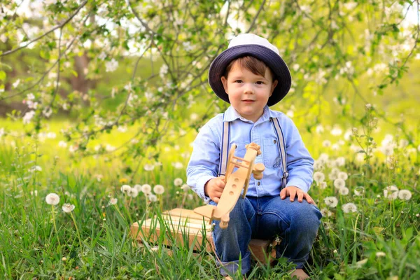 Kleiner süßer blonder Junge spielt an einem sonnigen Tag im Sommerpark auf dem Rasen mit einem Holzflugzeug — Stockfoto