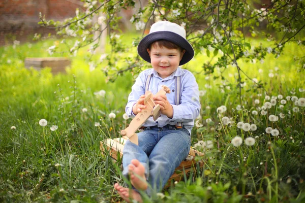Μικρό χαριτωμένο ξανθό αγόρι παίζει με ένα ξύλινο αεροπλάνο στο καλοκαιρινό πάρκο στο γρασίδι — Φωτογραφία Αρχείου