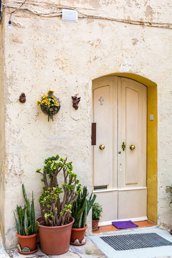 Vintage door in yellow limestone wall of old medieval building in Senglea, Malta, with angel figurines on the wall and potted plants on the floor. Senglea street view.