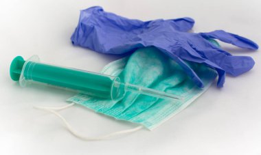 Tek kullanımlık yeşil maske, mavi lateks eldiven ve beyaz bir masada duran plastik şırınga. Ağız ve burnu kapamak için tipik cerrahi maske. Virüs koruma kavramı.