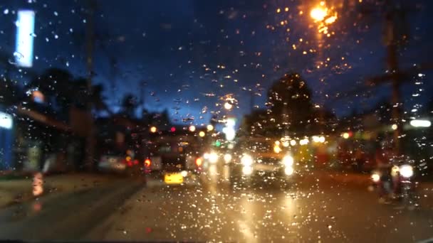 Tajski autobus z zapalonymi reflektorami podróżujący wzdłuż nocnej ulicy w porze deszczowej. Widok przez szybę samochodu w kroplach. Romantyczny widok typowej Azji wieczorem. Transport publiczny pod burzliwym niebem. — Wideo stockowe