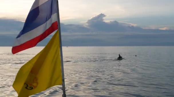Boot drijvend in de buurt van Thaise vlaggen in de avond. Silhouet van anonieme persoon drijvend op boot op kabbelende zee in de buurt van Thaise en Koning van Thailand vlaggen tegen bewolkte zonsondergang hemel. — Stockvideo