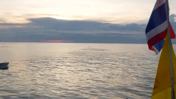 Boot schwimmt am Abend in der Nähe thailändischer Flaggen. Silhouette einer anonymen Person, die auf einem Boot auf dem plätschernden Meer in der Nähe thailändischer Flaggen schwimmt und König von Thailand vor wolkenverhangenem Himmel. — Stockvideo