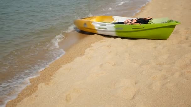 Каяк с веслами на песчаном берегу, омываемый волнами синего моря. Красочные каноэ в волнистой кристаллической воде, тропический одинокий остров пляж с копипространством. Активный отдых на свежем воздухе, летний отдых, спорт и здоровье — стоковое видео
