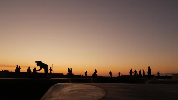 Silueta mladého skákajícího skateboardisty na dlouhém prkně, letní pozadí západu slunce. Venice Ocean Beach skatepark, Los Angeles California. Teenky na skateboardu, extrémní park. Skupina teenagerů. — Stock video