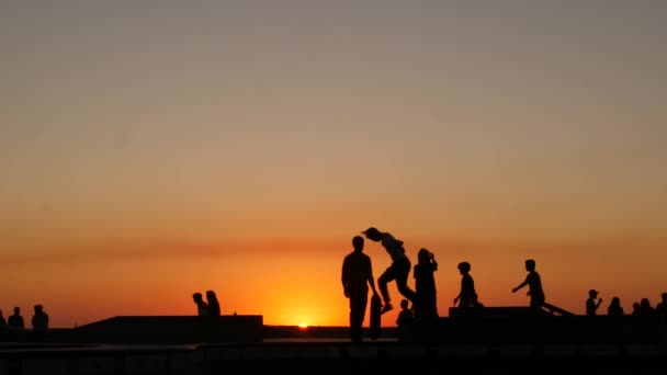 Sylwetka młodego skokowego deskorolkarza na longboardzie, letnie tło zachodu słońca. Skatepark Venice Ocean Beach w Los Angeles w Kalifornii. Nastolatki na rampie deskorolkowej, ekstremalny park. Grupa nastolatków. — Wideo stockowe