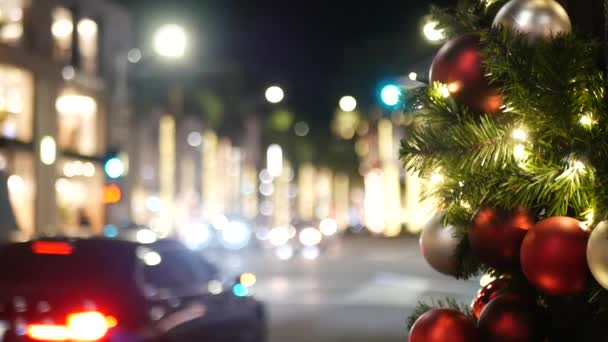 Primer plano del árbol de Navidad al aire libre decorado festivamente con bolas de color rojo brillante sobre el fondo borroso brillante hada. Luces de guirnalda desenfocadas, efecto Bokeh. Desenfocado calle de la ciudad de noche con coches en la carretera — Vídeo de stock