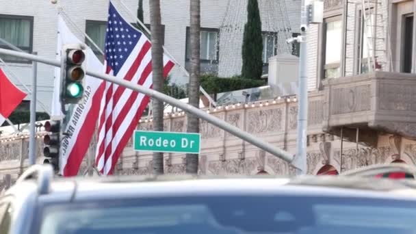 Всемирно известный знак Rodeo Drive Street Road в Беверли-Хиллз против американского флага Соединенных Штатов. Лос-Анджелес, Калифорния, США. Богатый потребитель жизни, роскошные бренды, концепция магазинов высокого класса. — стоковое видео