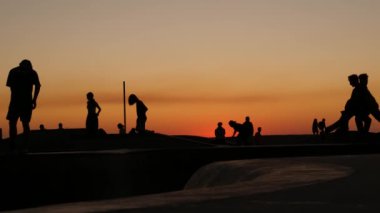 Yaz günbatımı arka planında, kaykayla gezen genç kaykaycı silueti. Venice Ocean Beach kaykay parkı, Los Angeles California. Kaykay rampasında gençler, ekstrem park. Bir grup genç..