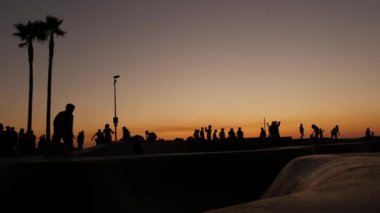 Yaz günbatımı arka planında, kaykayla gezen genç kaykaycı silueti. Venice Ocean Beach kaykay parkı, Los Angeles California. Kaykay rampasında gençler, ekstrem park. Bir grup genç..