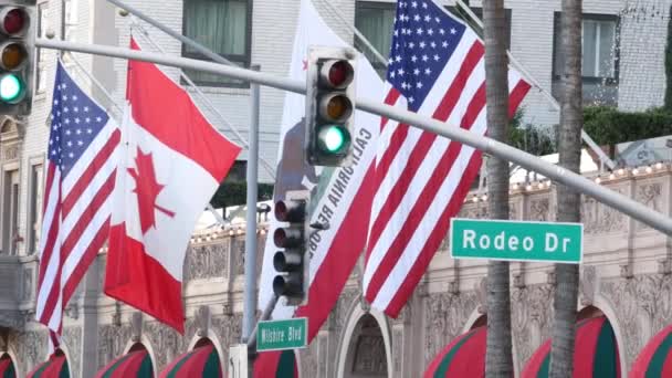 A világhírű Rodeo Drive Street Road Sign in Beverly Hills az Amerikai Egyesült Államok zászlaja ellen. Los Angeles, Kalifornia, USA. Gazdag gazdag élet fogyasztás, Luxus márkák, high-class üzletek koncepció. — Stock videók