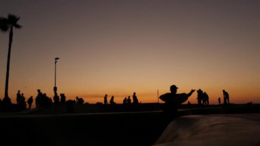 Yaz günbatımı arka planında, kaykayla gezen genç kaykaycı silueti. Venice Ocean Beach kaykay parkı, Los Angeles California. Kaykay rampasında gençler, ekstrem park. Bir grup genç..