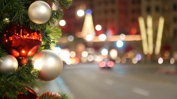 Nahaufnahme eines festlich geschmückten Weihnachtsbaums im Freien mit leuchtend roten Kugeln auf unscharfem, glitzerndem Feenhintergrund. Defokussierte Girlanden, Bokeh-Effekt. Entschärfte nächtliche Stadtstraße mit Autos auf der Straße — Stockvideo