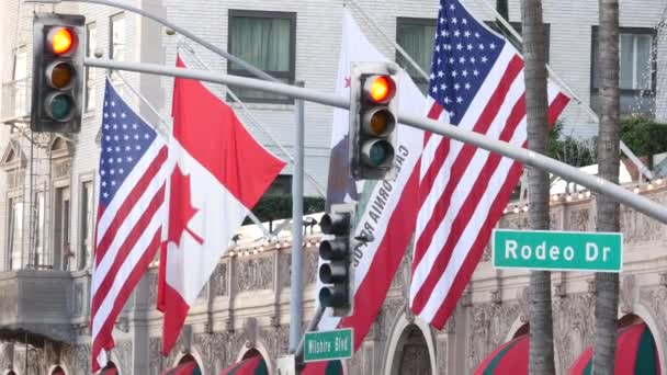 Señal de Rodeo Drive Street Road en Beverly Hills contra la bandera de Estados Unidos. Los Ángeles, California, EE.UU. Consumismo rico de la vida, marcas de lujo, concepto de tiendas de clase alta. — Vídeo de stock