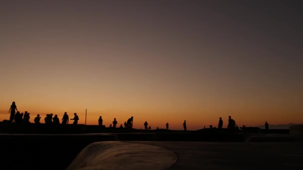 Sylwetka młodego skokowego deskorolkarza na longboardzie, letnie tło zachodu słońca. Skatepark Venice Ocean Beach w Los Angeles w Kalifornii. Nastolatki na rampie deskorolkowej, ekstremalny park. Grupa nastolatków. — Wideo stockowe
