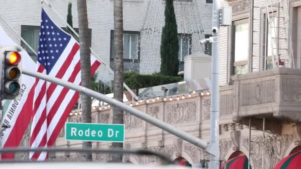 A világhírű Rodeo Drive Street Road Sign in Beverly Hills az Amerikai Egyesült Államok zászlaja ellen. Los Angeles, Kalifornia, USA. Gazdag gazdag élet fogyasztás, Luxus márkák, high-class üzletek koncepció. — Stock videók