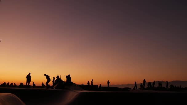 Silhouette fiatal ugró gördeszka lovaglás longboard, nyári naplemente háttér. Venice Ocean Beach skatepark, Los Angeles Kalifornia. Tinik gördeszkás rámpán, extrém park. Tinédzserek csoportja. — Stock videók