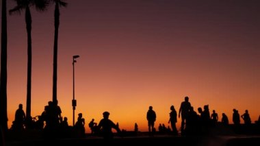 Yaz günbatımı arka planında, kaykayla gezen genç kaykaycı silueti. Venice Ocean Beach kaykay parkı, Los Angeles California. Kaykay rampasında gençler, ekstrem park. Bir grup genç..