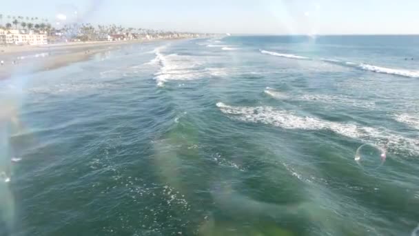 Fukující mýdlové bubliny na mořském molu v Kalifornii, rozmazané letní pozadí. Tvůrčí romantická metafora, koncept snění o štěstí a magii. Abstraktní symbol dětství, fantazie, svobody — Stock video