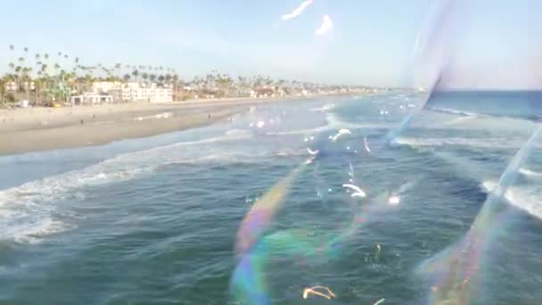 Вибух мильних бульбашок на океанському пірсі в Каліфорнії, розмитий літній фон. Творча романтична метафора, концепція мрії щастя і магії. Абстрактний символ дитинства, фантазії, свободи — стокове відео
