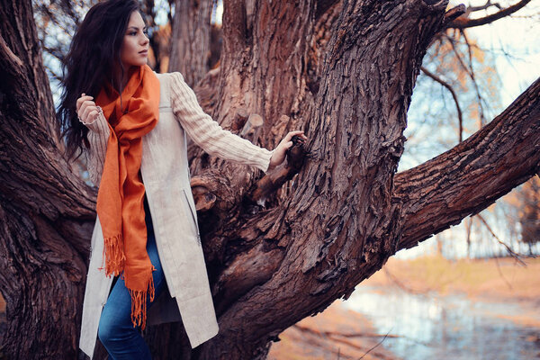 Young beautiful woman with orange scarf posing against big tree in autumn park