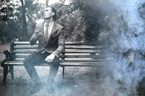 公園のベンチに座っていると スマート フォンを使用してのスーツでハンサムな若い新郎 — ストック写真
