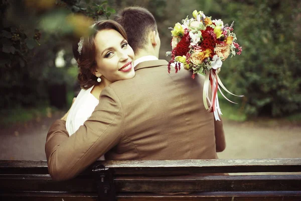 花とハンサムな新郎の結婚式の日に公園のベンチに座っての花束と美しい若い花嫁 — ストック写真