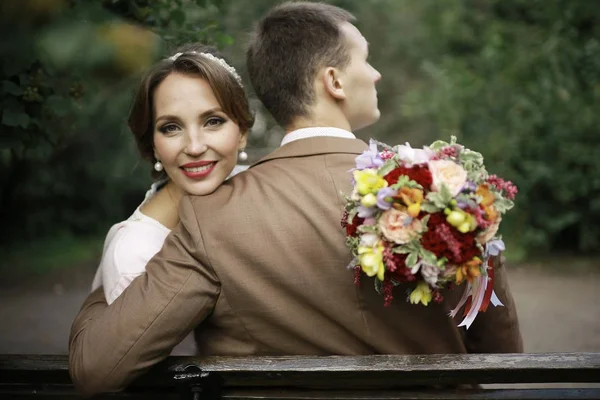 花とハンサムな新郎の結婚式の日に公園のベンチに座っての花束と美しい若い花嫁 — ストック写真