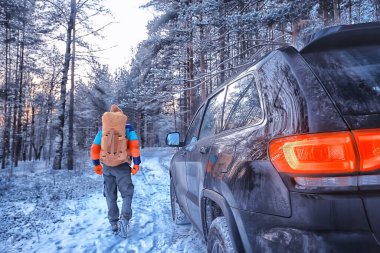 Orman manzaralı bir arabanın yanında sırt çantalı bir erkek gezgin kış yürüyüşü yapıyor.