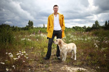 İtaatkar Labrador Retriever köpeğini eğiten ve bulutlu yaz gününde yeşil çiçek çayırında temel komutları veren genç bir adamın yan görüntüsü.