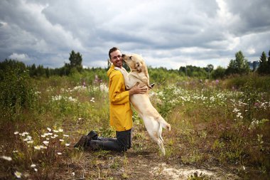Sarı ceketli neşeli bir erkeğin önünde diz çöküp yetişkin Labrador Retriever 'ı kucaklarken mutlu köpek sahibinin yüzünü yalıyor.
