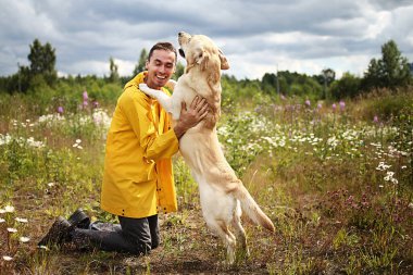 Sarı ceketli neşeli bir erkeğin önünde diz çöküp yetişkin Labrador Retriever 'ı kucaklarken mutlu köpek sahibinin yüzünü yalıyor.