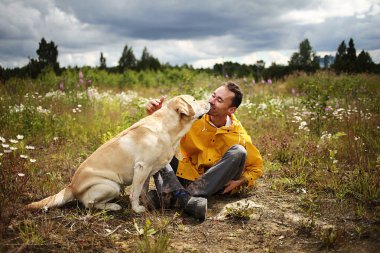 Arka planda bulutlu gökyüzü olan çiçek açan çayırda bej renkli Labrador av köpeğiyle öpüşen genç adamın yan görüntüsü