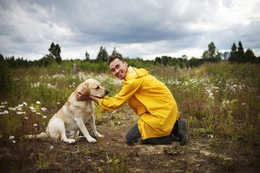 Labrador Retriever 'ın önünde tek dizinin üstünde dururken gülümseyerek gülen rahat genç bir adamın yan görüntüsü